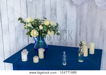 white flowers roses in a blue vase on a blue table with a white wooden background.