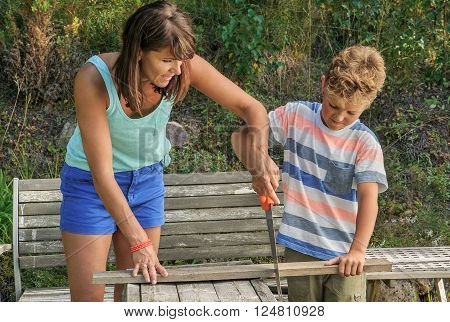 Mother teaches son how to use a saw. They're outside in the garden. Traditional man's role played by woman