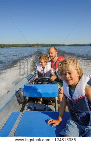 Father and sons out boating together. The father lets one of the boys steer the boat. They're wearing life vests. In the background there's forested shore line water and blue sky.