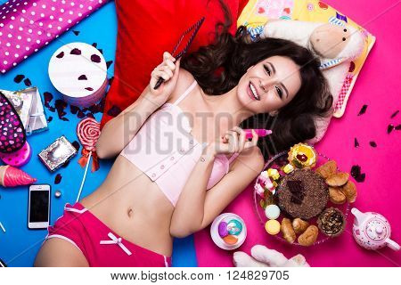 Beautiful fresh girl doll lying on bright backgrounds surrounded by sweets, cosmetics and gifts. Fashion beauty style. Photos shot in the studio.