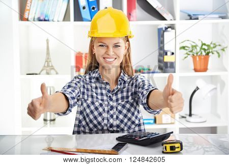 Young prosperous woman engineer in hardhat showing thumbs up at her workplace. Attractive female architect measuring blueprint and calculating business plan.
