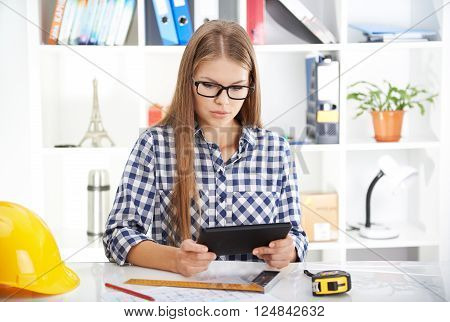 Concentrated female architect with digital tablet studying construction plans. Young woman designer sitting at table with computer and engineering tools.