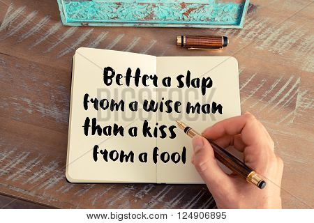 Retro effect and toned image of a woman hand writing on a notebook. Handwritten quote Better a slap from a wise man than a kiss from a fool as inspirational concept image