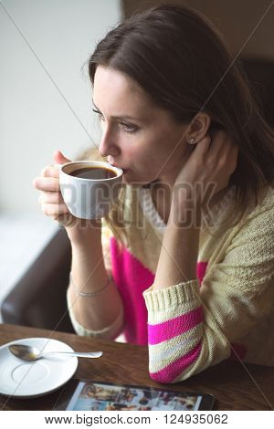 leisure, drinks, people, technology and lifestyle concept - young woman drinking coffee in a cafe in the trendy sweater, looking out the window uses the tablet waits in social networks, online