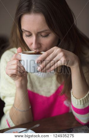 Young happy girl drinking hot coffee or tea sits in a cafe, lifestyle, breakfast or lunch enjoys relaxing, healthy lifestyle, fashion, in a yellow sweater.