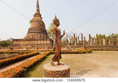 Walking Buddha statue past Buddhist stupa inside Sukhothai historical park Thailand. UNESCO World Heritage Site.