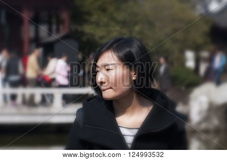 A chinese woman looking off to her right as she is thinking outside in a classic chinese garden.