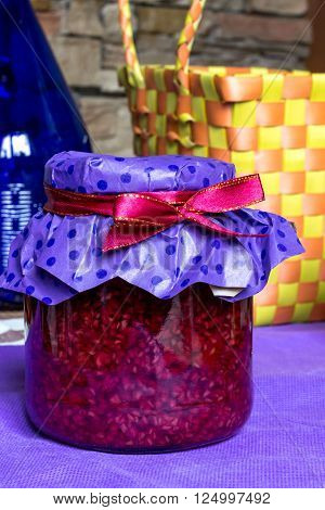 closeup photo of sweet raspberry jam-jar, with red  ribbon and craft paper, strow busket, blue bottle on table in kitchen, food photography