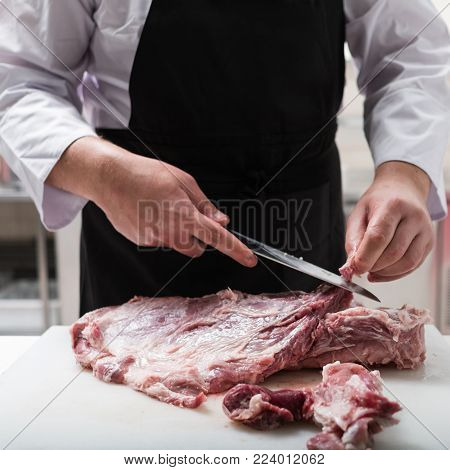 Restaurant chef in black apron cutting fresh lamb meat in the kitchen. Ingredient preparation before cooking a delicious meal