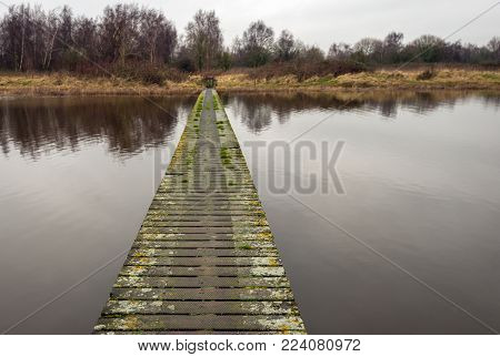Long wooden bridge over the water of a wide creek at a nature reserve. The planks of the bridge are weathered and covered with mosses and lichens. To prevent slipping, the bridge deck is covered with wire mesh.