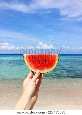 Hand holding slice of watermelon on the beach. Sea and blue sky background. Summer mood.