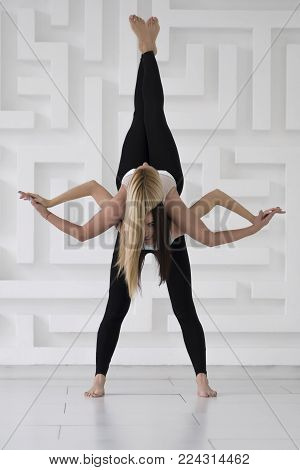 Two young girls practicing acro yoga balance pose with legs up and crossed arms, black sportswear, white abstract background, studio shoot