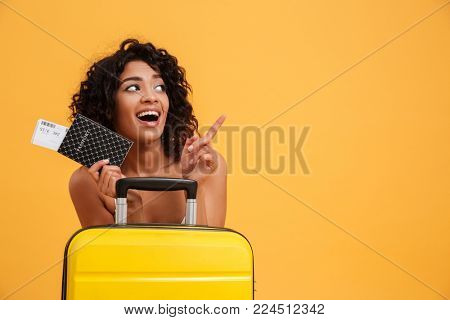 Portrait of a cheerful young african woman dressed in summer clothes holding passport with flying tickets while standing with a suitcase and pointing finger away isolated over yellow background