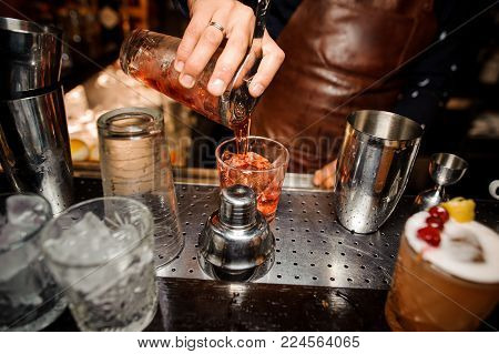 barman in a brown apron is pouring out of a shaker into a crystal glass an alcoholic cocktail of amber color, against a bar counter, glasses with ice and ready-made cocktails