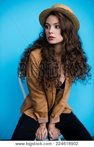 seductive young woman in canotier hat and jacket sitting on chair and looking away