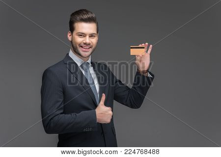 smiling businessman holding golden credit card and showing thumb up, isolated on grey