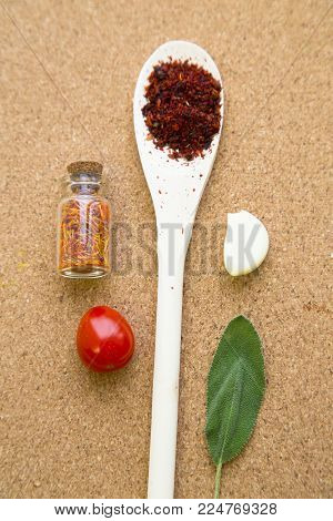 Cooking hot spicy meal. A wooden spoon with crushed chili pepper, dry herbs in a glass jar, fresh cherry tomato, a salvia leaf and a garlci cloves, corkwood background, top view