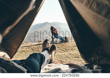 Woke up in the perfect place. Unrecognizable young man enjoying the view of mountain range from the tent while his girlfriend resting near the campfire