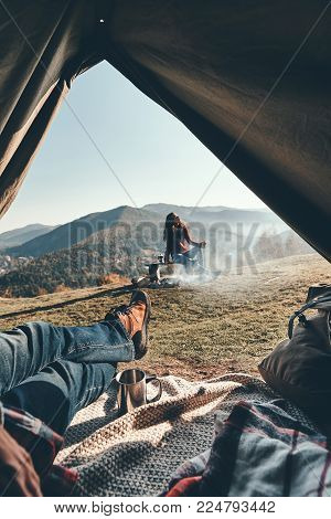 Unbelievable beauty. Unrecognizable young man enjoying the view of mountain range from the tent while his girlfriend resting near the campfire