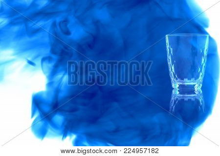 Faceted shot empty shot of vodka or tequila on white background enveloped in puff of blue smoke. Place for your text.