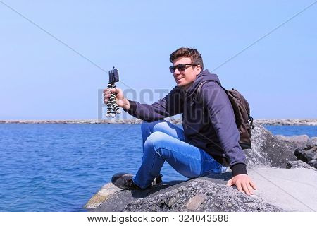Traveller Man Talks Video Chat On Smartphone With Small Tripod Comes To Sea Beach Sits On Stone. Tou