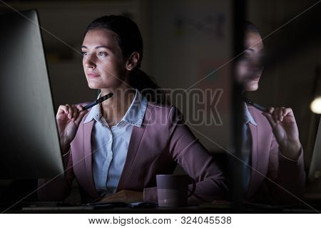 Concentrated Young Manager Looking At Computer Monitor While Working At Her Workplace Till Night At 