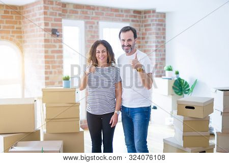 Middle age senior couple moving to a new home with boxes around doing happy thumbs up gesture with hand. Approving expression looking at the camera with showing success.