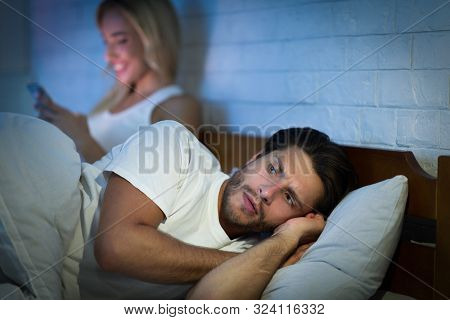 Jealousy And Cheating. Girlfriend Texting On Mobile Phone Lying With Boyfriend In Bed At Night. Low 