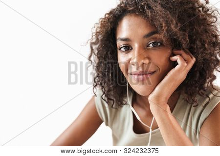 Smiling young african woman listening to music with earphones while lening on her hand and looking at camera isolated over white background