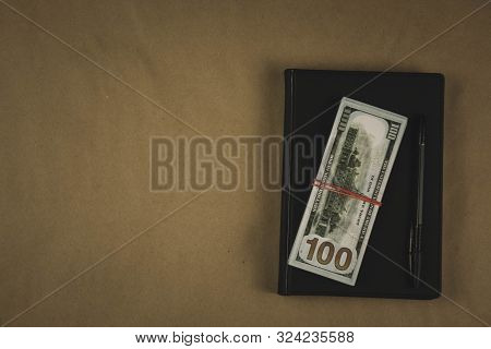 A Black Notebook , A Pen And Dollar Cash Banknotes On Wooden Background - Concept Of Financial Manag
