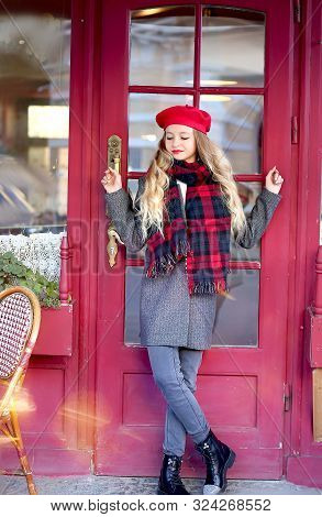 Girl In A Gray Coat And A Red Beret With Long Curls Stands Thoughtfully Near A Cafe On A Background 