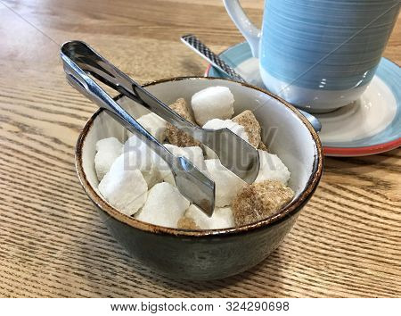 Bowl With Pieces Of White And Brown Sugar Cubes And With Sugar-tongs On A Wooden Table In Front Of A