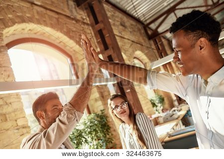 Celebrating Success. Two Cheerful Men Giving High-five While Standing With Colleagues In The Modern 