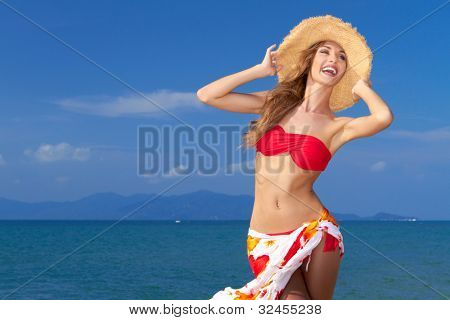 Glamourøse leende curvaceous kvinde i bikini og solhat poserer foran havet med hendes lange 