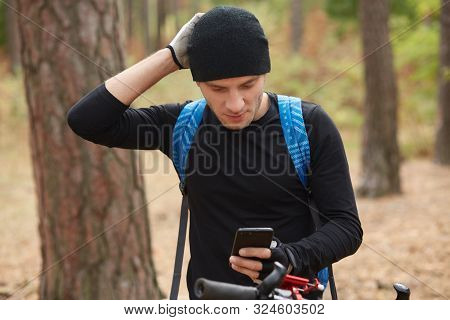 Handsome Young European Cyclist Wearing Black Sportwear And Blue Bacpack, Messaging Friends On Mobil