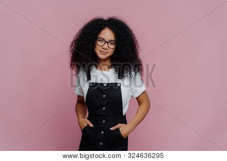 Portrait Of Happy Curly African American Woman Keeps Hands In Pockets Of Sarafan, Wears Transparent 