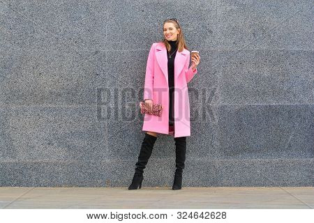 Beautiful And Happy Woman In Pink Coat  And Knee-high Boots Standing With Coffee Cup Outdoors