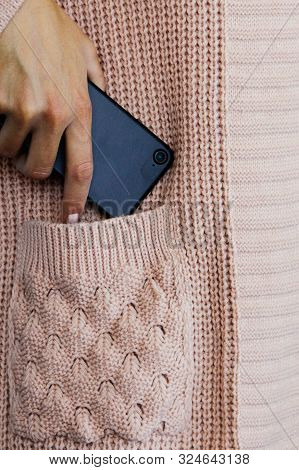 Element Of A Female Knitted Cardigan. A Female Hand Lays A Black Mobile Phone In The Pocket Of A Kni