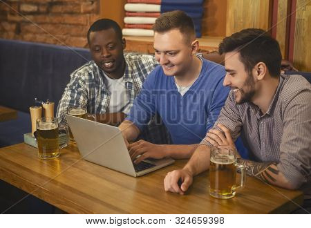 Dating Website. Three Happy Young Men Drinking Beer And Using Lapop In Pub Together