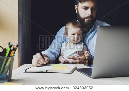 Father With Little Baby On Knees Making Notes In Notebook, Looking At Laptop. Young Man Holding Chil