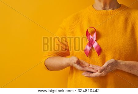 Senior woman on color yellow background. Pink ribbon like a symbol of breast cancer awareness. Support people living with tumor illness.