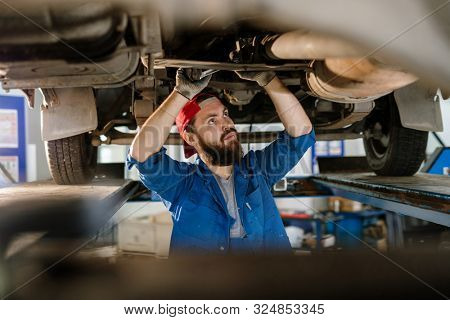 Serious worker in workwear standing under broken car, examining its motor and trying to restrain the problem