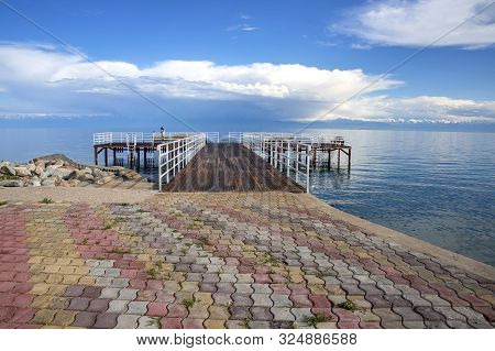 Pier On Issyk-kul Lake On The Background Of A Mountain Range With Snowy Peaks And Cloudy Sky. Kyrgyz