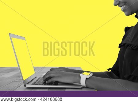 Composition of smiling woman wearing smartwatch sitting at table using laptop, on bright yellow. global business, communication and digital interface concept digitally generated image.
