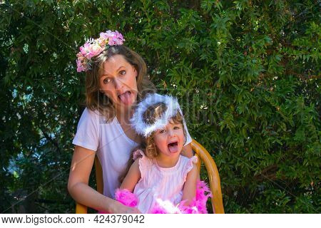 Happy beautiful young mother in a flower wreath and adorable little daughter laugh. Background dense evergreen hedge. Concept - portrait and advertising photo