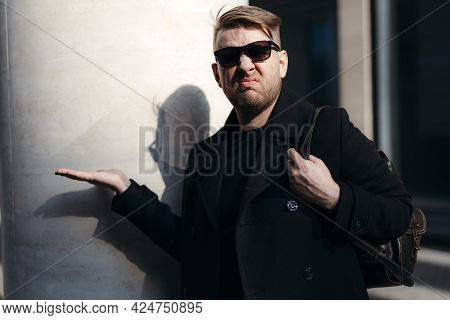 Handsome Young Guy In A Black Coat In Sunglasses And With A Backpack On His Shoulder Looks Displease