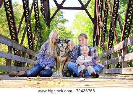 Family Of Four People And Dog Sitting On Bridge In Autumn