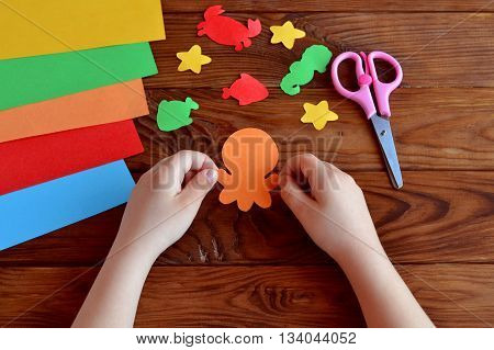 Cut out of colored paper marine animals - octopus, fish, starfish, seahorse, crab. Little child holds a octopus crafts in hands. Kids diy, sheets of colored paper, scissors on wooden background