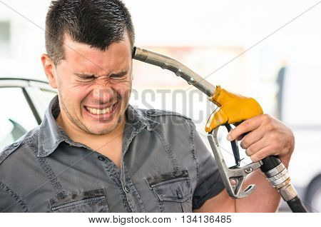 Young man in suicide mimic position at fuel gas station - Gasoline price rising concept with desperate guy pointing pump nozzles pistol on head - Shallow depth of field with focus on closed eyes