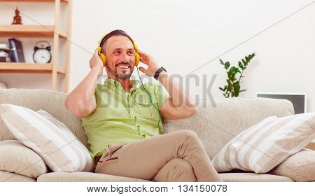 Picture of handsome man resting and relaxig while sitting on sofa or couch listening to music at home. Happy man touching his earphones.